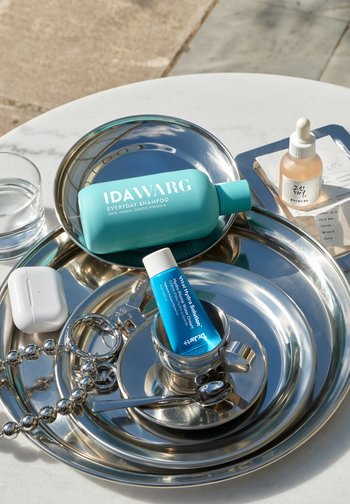 Sininen IDAWARG-shampoopullo ja sininen Dr. Jart+ -voidepullo kiiltävillä metallialustoilla valkoisen AirPods-kotelon ja pipettipullon kanssa valkoisella pöydällä.