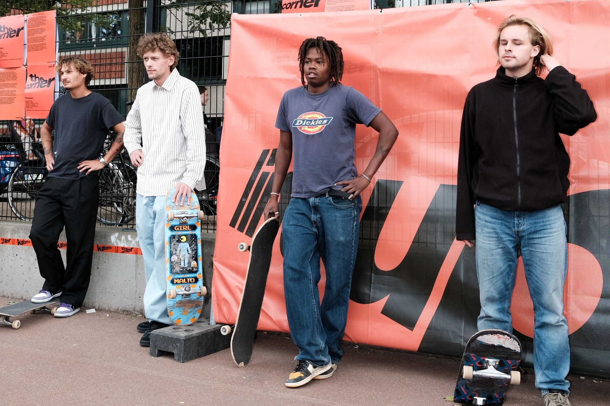 Fyra personer står mot en orange bakgrund. Två håller skateboards och är klädda i avslappnade kläder: en i en blå skjorta och den andra i en randig skjorta.