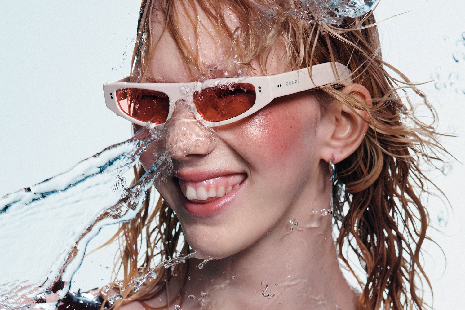 A young person with wet, wavy hair smiles playfully, wearing white sunglasses with orange lenses, as water splashes around them.