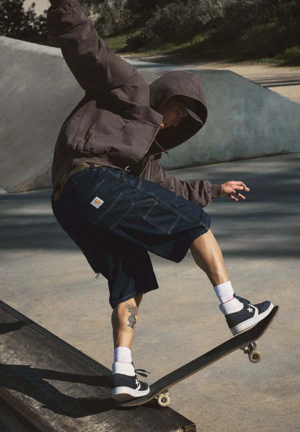 Skateboarder wearing a hoodie and shorts performs a trick on a ramp at an outdoor skatepark with trees in the background.