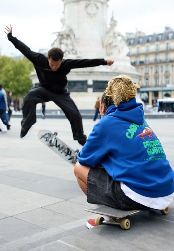 Två skateboardåkare i ett urbant torg. En gör ett trick i svart sportklädsel, medan en annan sitter på en skateboard i en blå hoodie och shorts.
