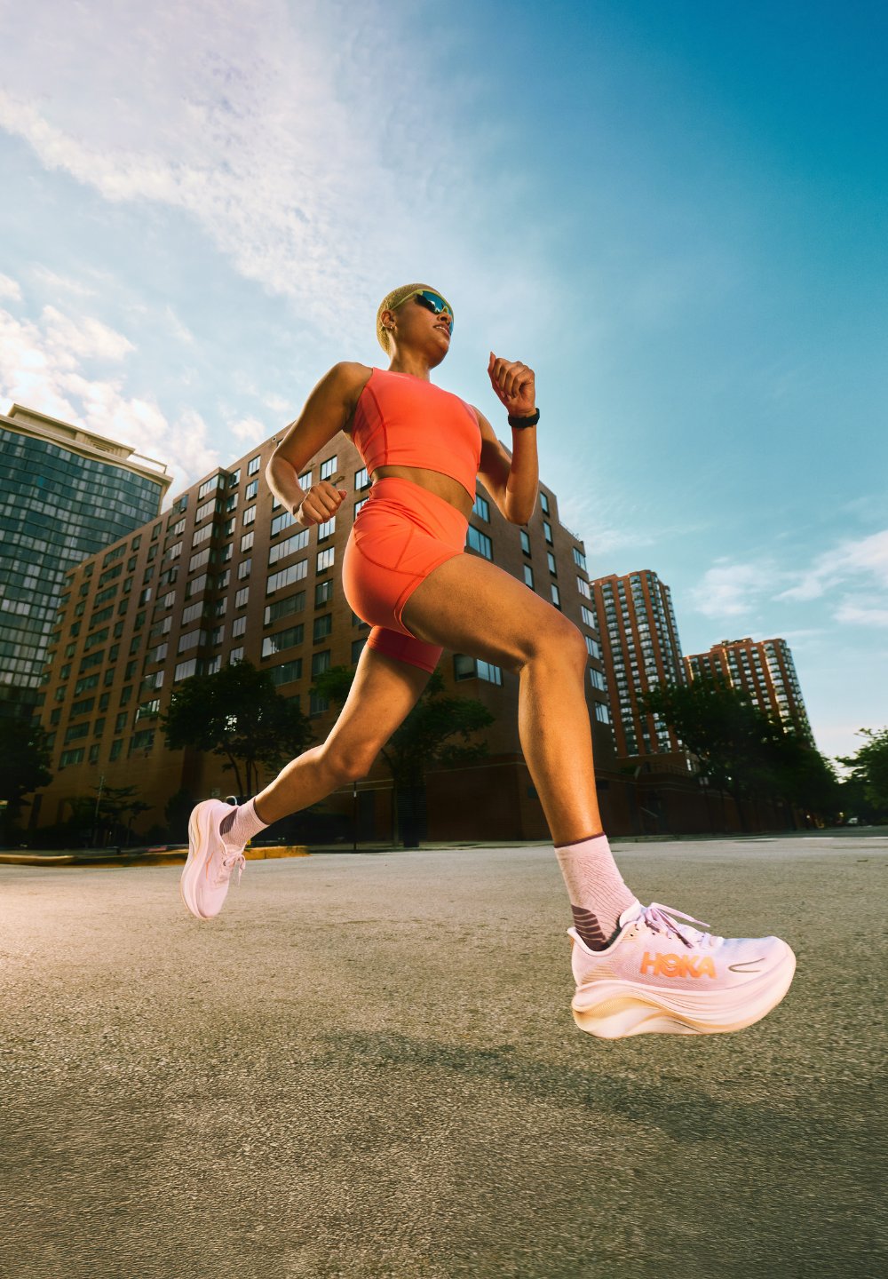 Coureur en tenue de sport orange et chaussures blanches sprintant dans une rue urbaine avec de hauts bâtiments et un ciel bleu en arrière-plan.