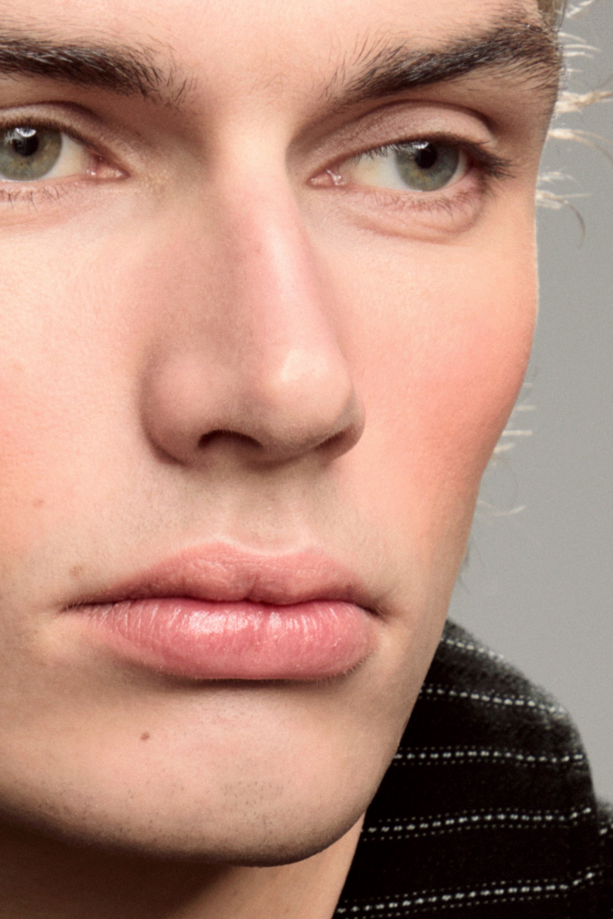 Close-up of a young man's face, highlighting his striking features, smooth skin, and neutral expression against a blurred gray background.
