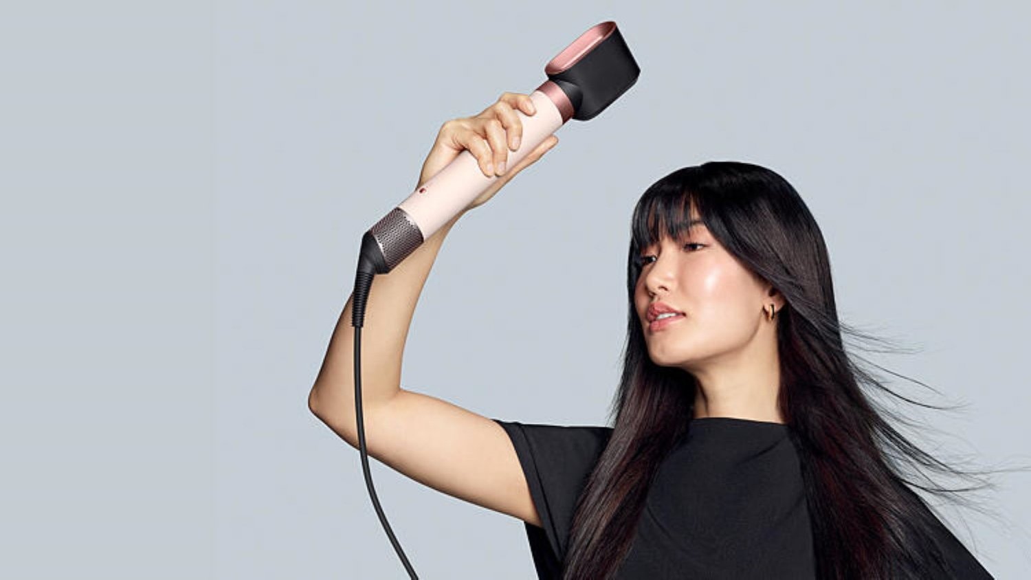 Frau mit langen dunklen Haaren, die einen rosa-schwarzen Haartrockner benutzt und die Haare nach hinten bläst vor einem hellgrauen Hintergrund.