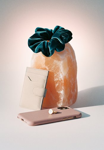 Elastic verde din catifea pe un lampă de sare Himalaya, suport pentru card bej sprijinit de lampă, smartphone roz cu inel auriu pe o suprafață albă.