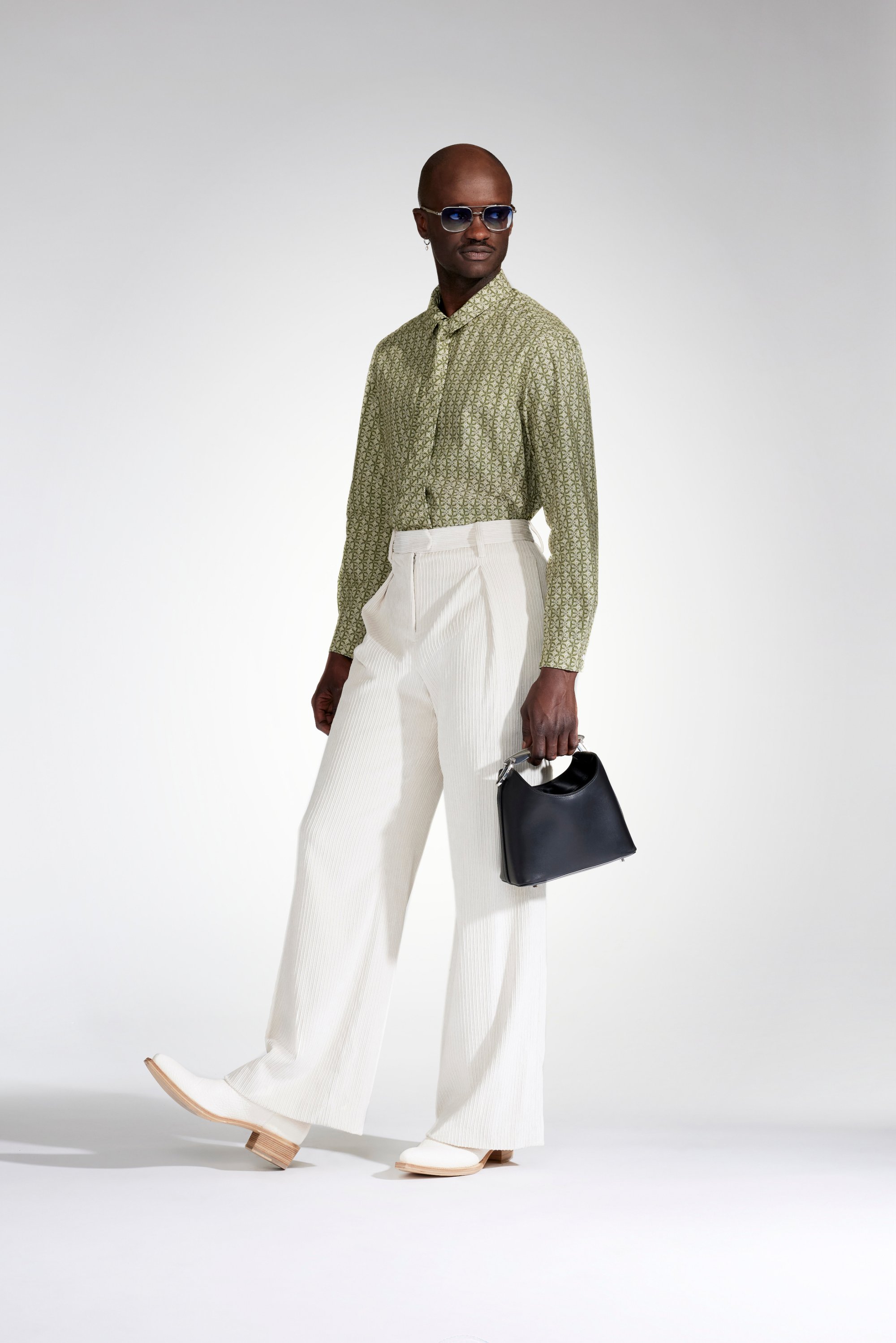 A model poses confidently in a patterned green shirt and white wide-legged trousers, holding a black handbag, wearing sunglasses.