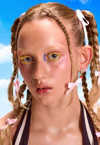 Une jeune personne avec des cheveux tressés ornés de rubans roses et de maquillage coloré pose devant un ciel bleu lumineux avec des nuages.