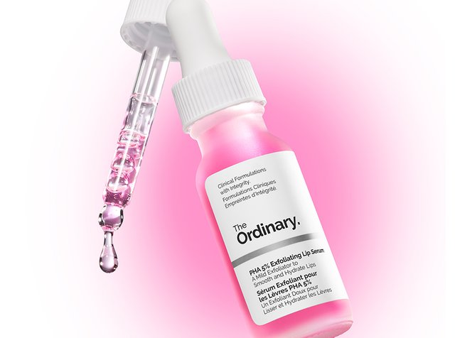 Botella de vidrio rosa con cuentagotas, etiquetada como "The Ordinary" suero exfoliante labial PHA 5%. Superficie lisa, tapa blanca y gota de líquido visible.