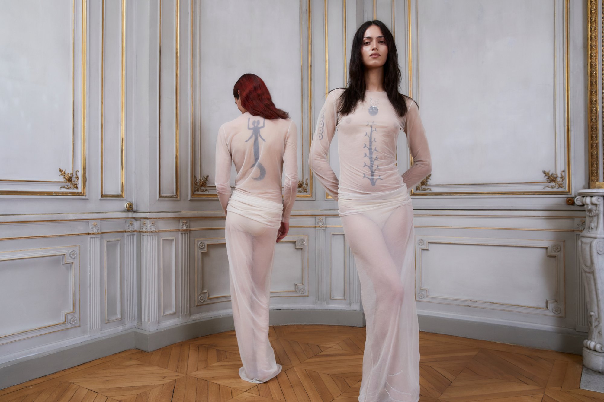 Due modelli in abiti trasparenti e aderenti con intricati tatuaggi corpo si trovano in una stanza elegante con dettagli ornamentali e pavimento in legno.