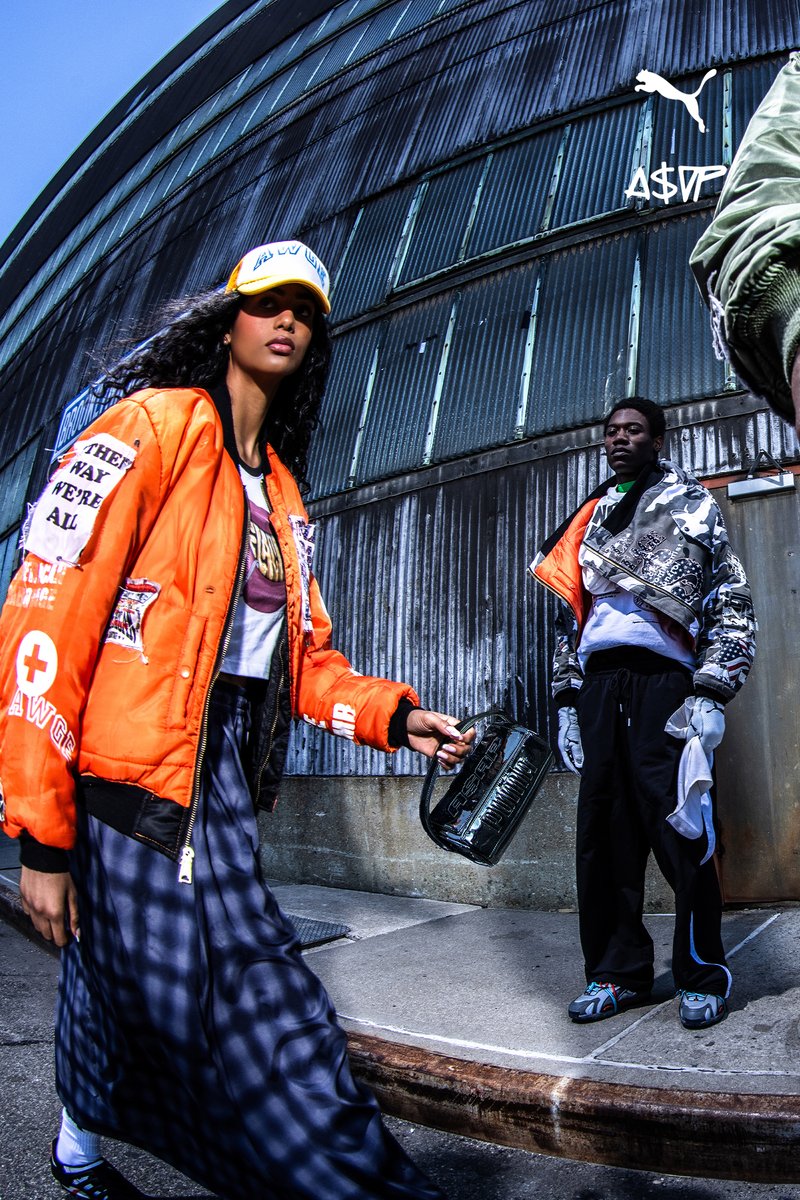 Two models pose against a textured metal backdrop. One wears an orange bomber jacket and holds a purse, while the other sports a patterned coat.