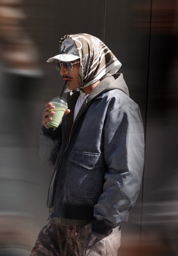 Homme portant des lunettes de soleil, un foulard à motifs sur une casquette, une veste grise et un pantalon camouflage, sirotant un smoothie vert avec une main dans sa poche.