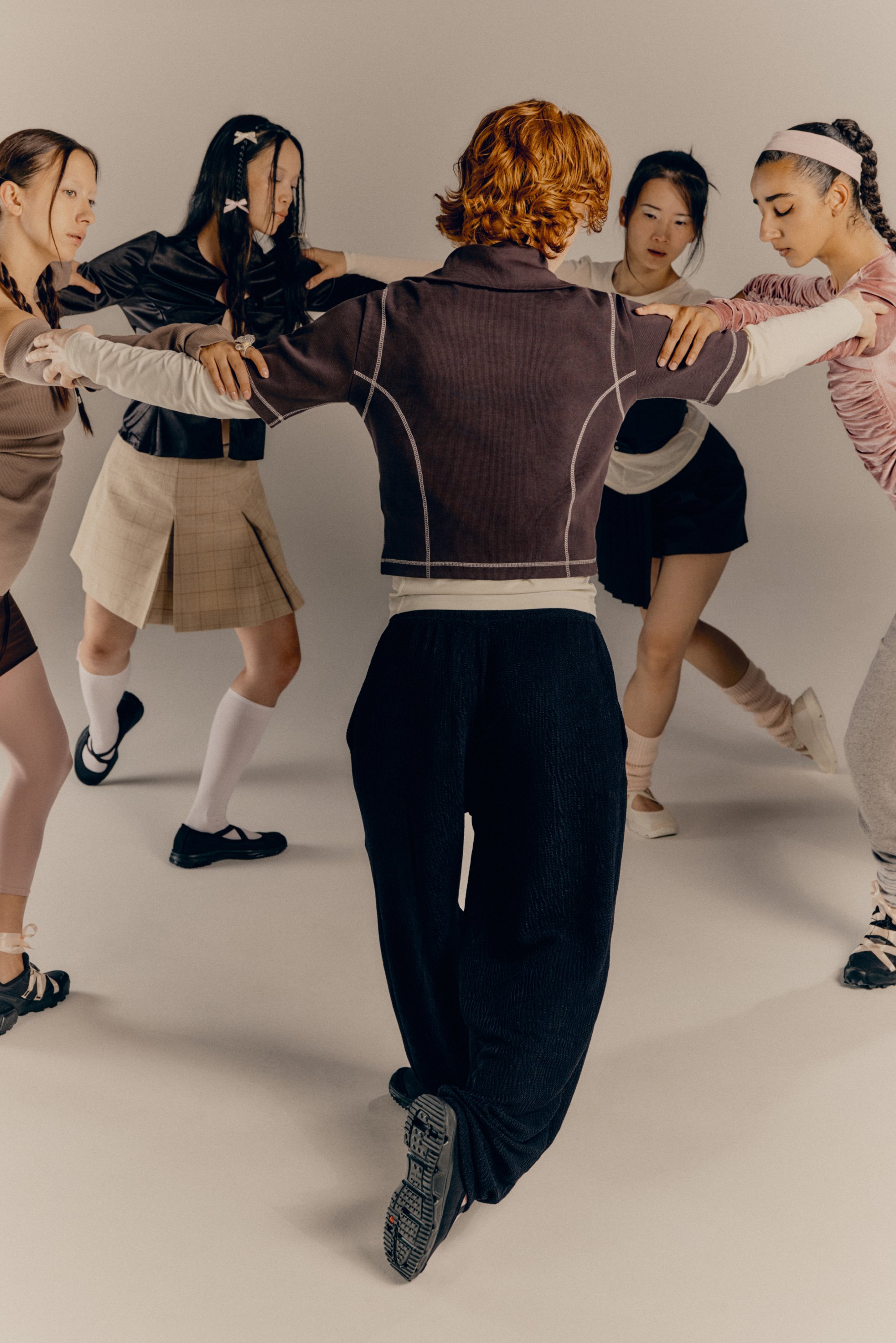 Vijf diverse individuen werken samen in een dansformatie, met elkaar verbonden door handen, gekleed in stijlvolle outfits op een neutrale achtergrond.