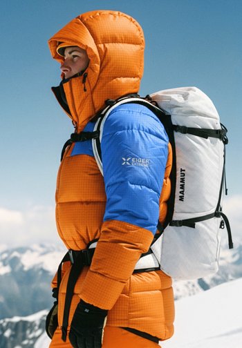 Oransje og blå dunjakke med rutenettmønster og hette, kombinert med en hvit ryggsekk med svarte detaljer og stropper.