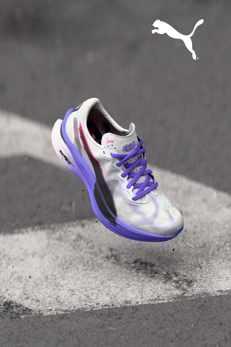 Zapatilla Puma Deviate con parte superior de malla blanca, acentos morados, panel lateral texturizado en negro y suela gruesa de color morado y rosa, levitando por encima del suelo.