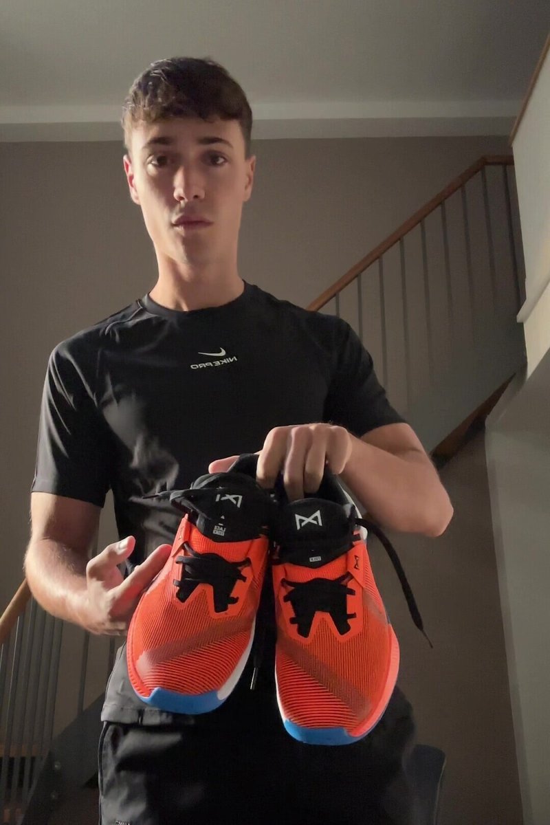 Jasně oranžové sportovní boty s černými doplňky, strukturovaným síťovaným svrškem, tkaničkami a modrou podrážkou, držené osobou v černém tričku.