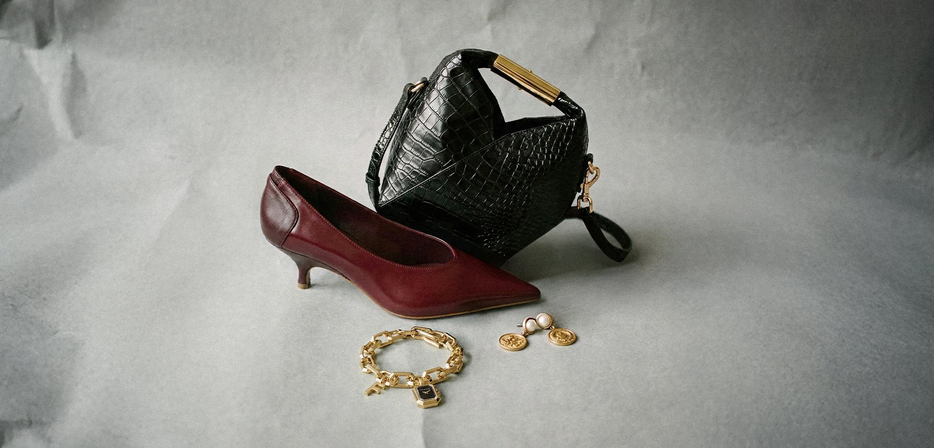Burgundy leather high-heeled shoe, black textured handbag, gold chain bracelet, and round earrings with pearl accents on grey background.
