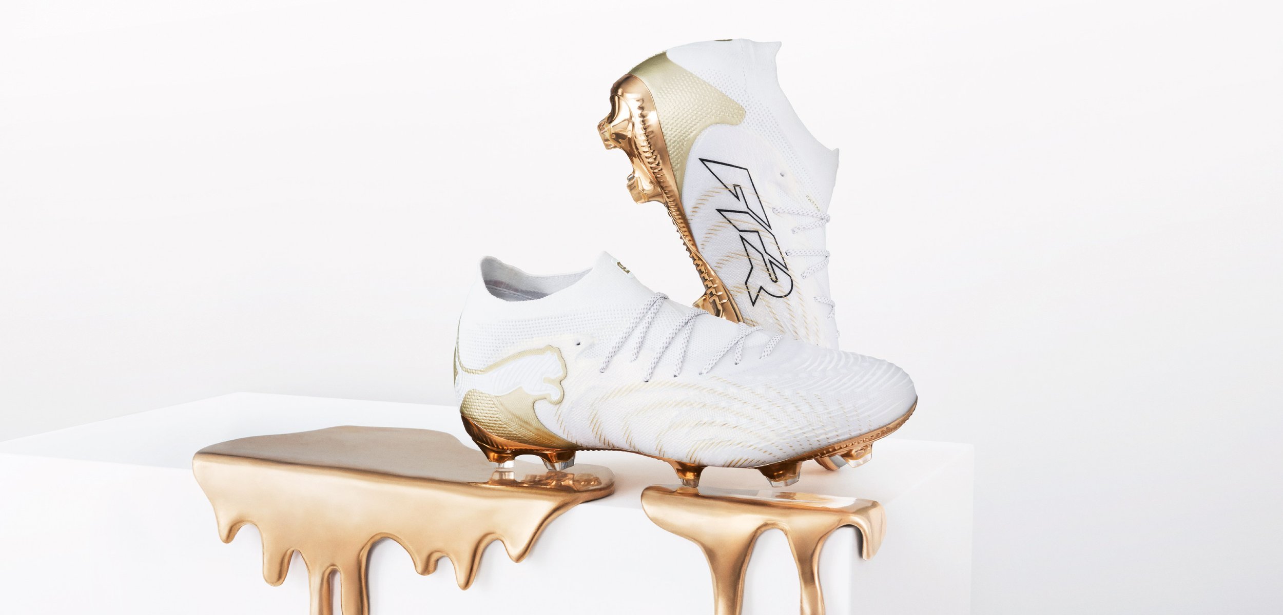 Par hvite fotballsko med gullsåler og detaljer, vist på en overflate med gullvæske som drypper over kantene.