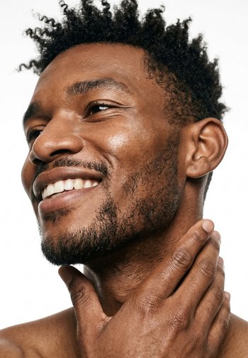 Smilende mand med krøllet hår, der blidt rører sin nakke og viser klar hud og ansigtshår mod en ensfarvet hvid baggrund.