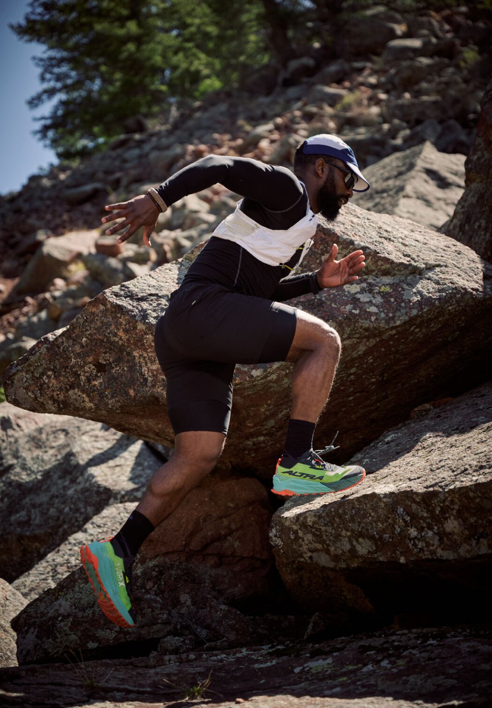 Silhouette athlétique en short noir et débardeur blanc, sautant par-dessus des rochers. Porte une casquette marine et des chaussures de course vertes avec des accents orange.