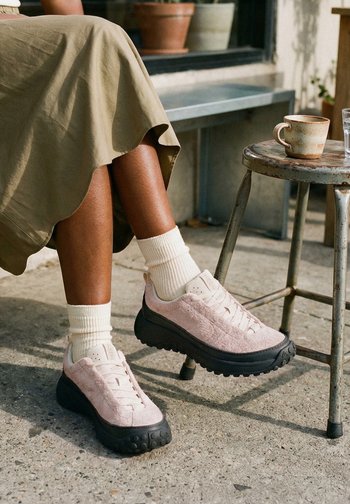 Persona con falda beige, calcetines blancos y zapatillas rosas sentada junto a una mesa rústica de metal sosteniendo una taza de cerámica y un vaso de agua.