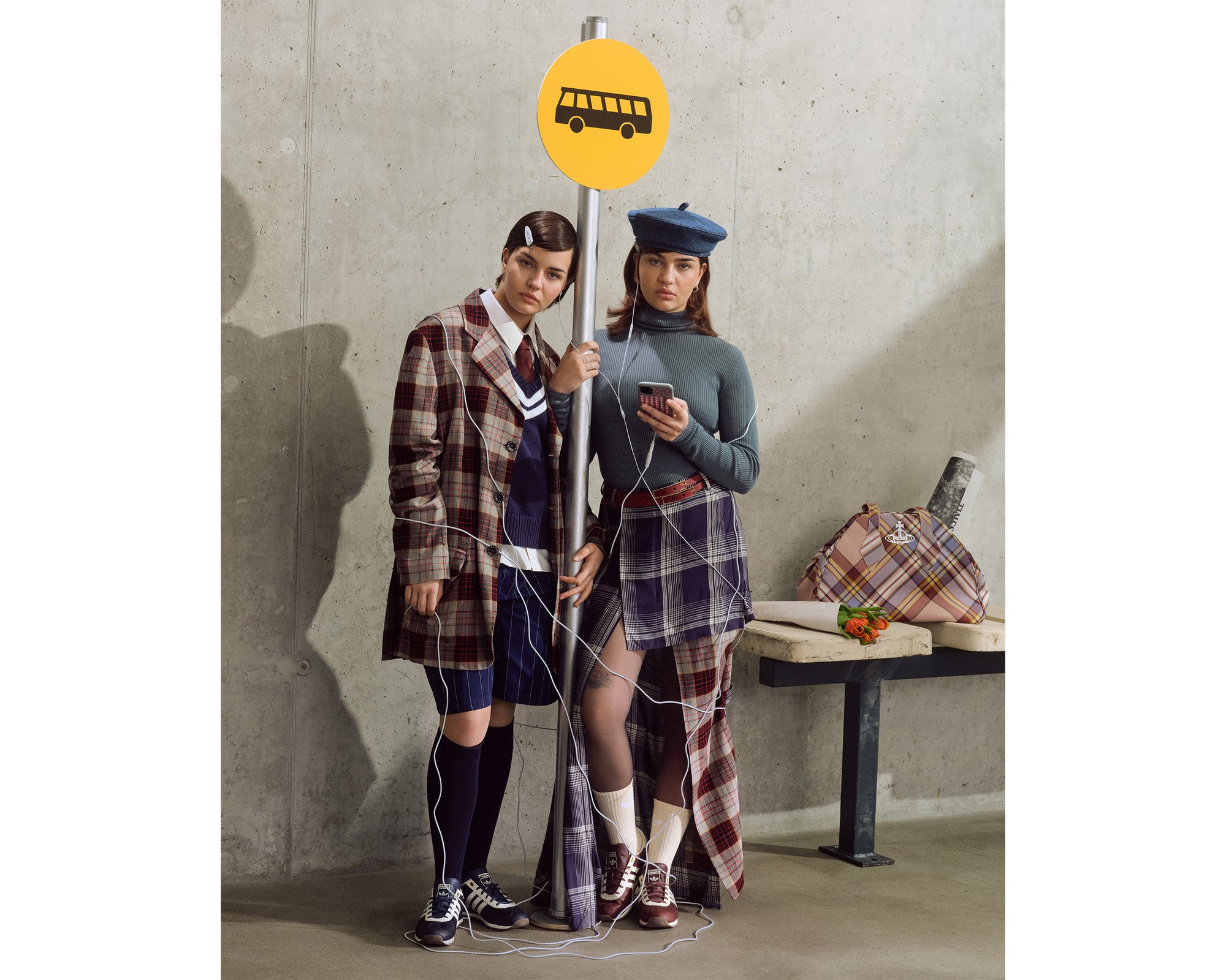 Twee modellen staan bij een bushaltebord, gekleed in geblokte jassen, rokken en accessoires. Een bruine handtas ligt op een bank.