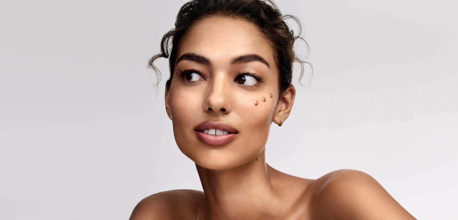 Young woman with clear skin and three skincare product drops on her cheek looking to the side against a plain background.