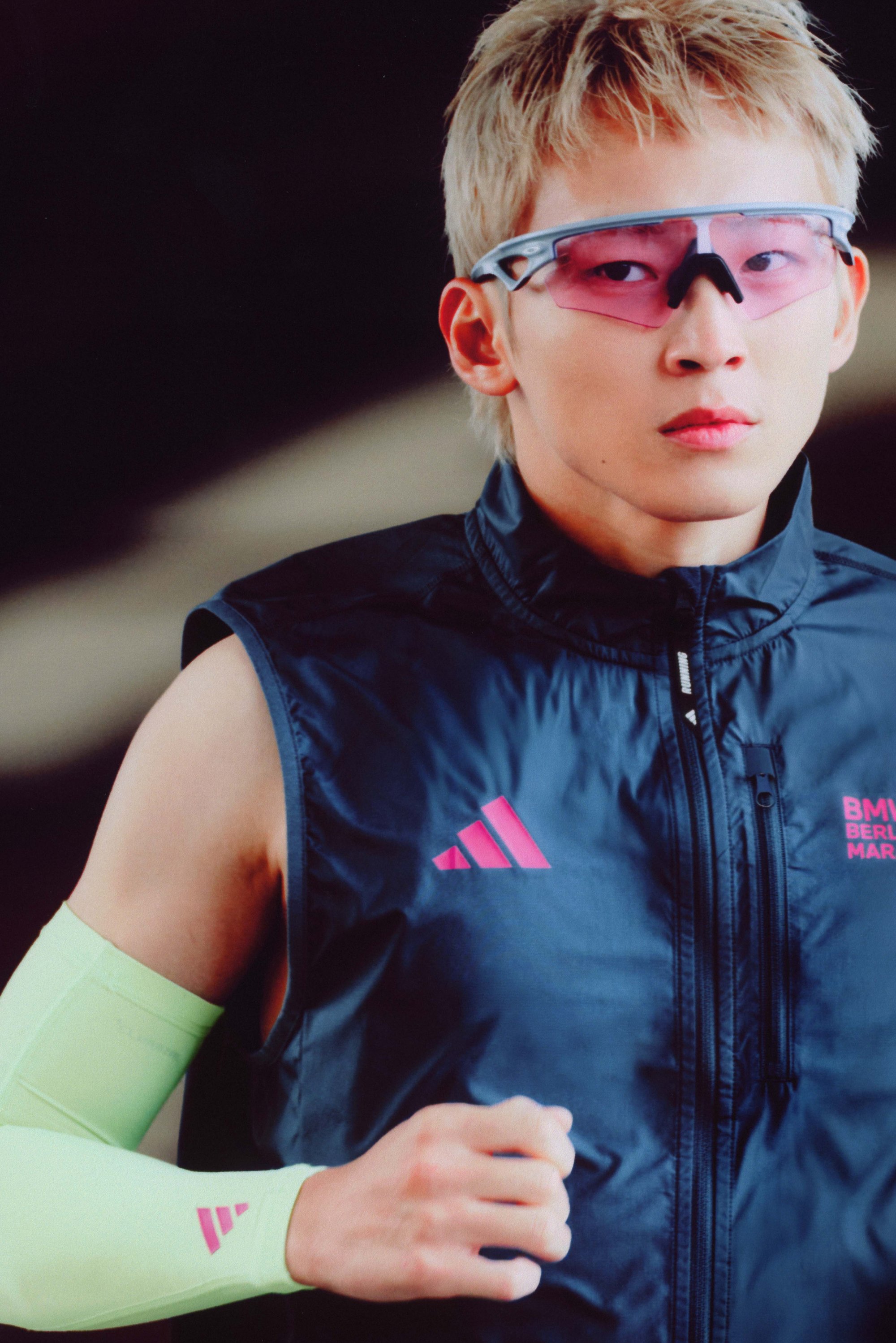 Athlete wearing a black sleeveless jacket with pink accents and a neon green arm sleeve. The jacket has a zip and logo detail.