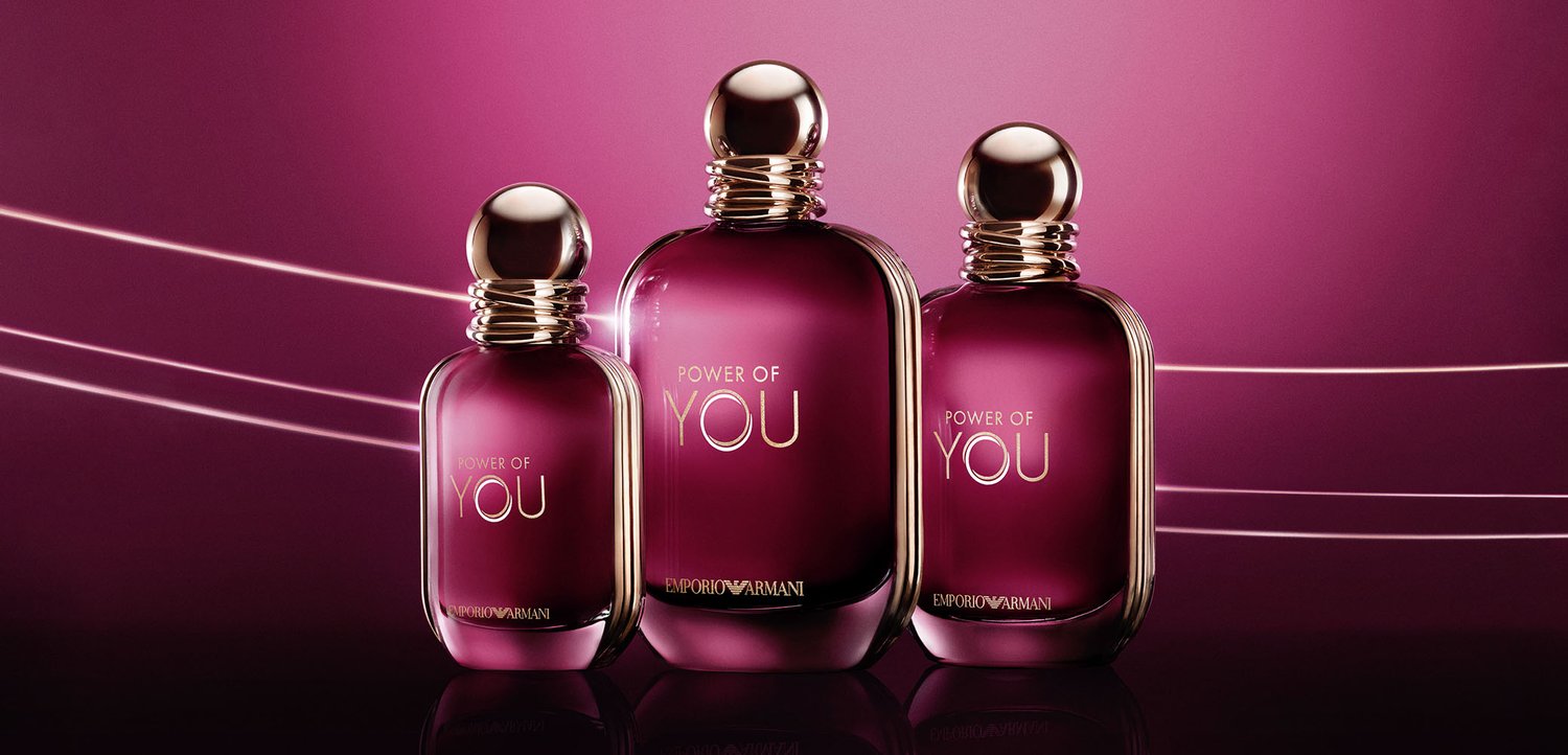 Drei lila Glasparfümflaschen mit goldenen Verschlüssen, auf denen "Power of You" von Emporio Armani steht, auf einer reflektierenden Oberfläche vor einem lila Hintergrund.