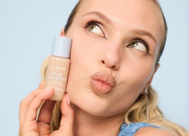 Vrouw met naar achteren gebonden haar houdt een klein flesje met het etiket "Lumene Blur Even Cover" dicht bij haar gezicht, terwijl ze omhoog kijkt met getuite lippen.