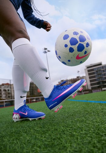 Persona, kas valkā zilus un rozā futbola apavus un baltas zeķes, sit baltu un zilu futbola bumbu uz zālaina laukuma.