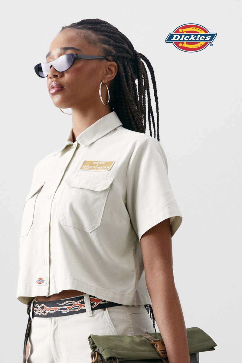 Een vrouw met gevlochten haar draagt een oversized zonnebril en een lichtgekleurde shirt met Dickies en Harley-Davidson logo's, tegen een neutrale achtergrond.