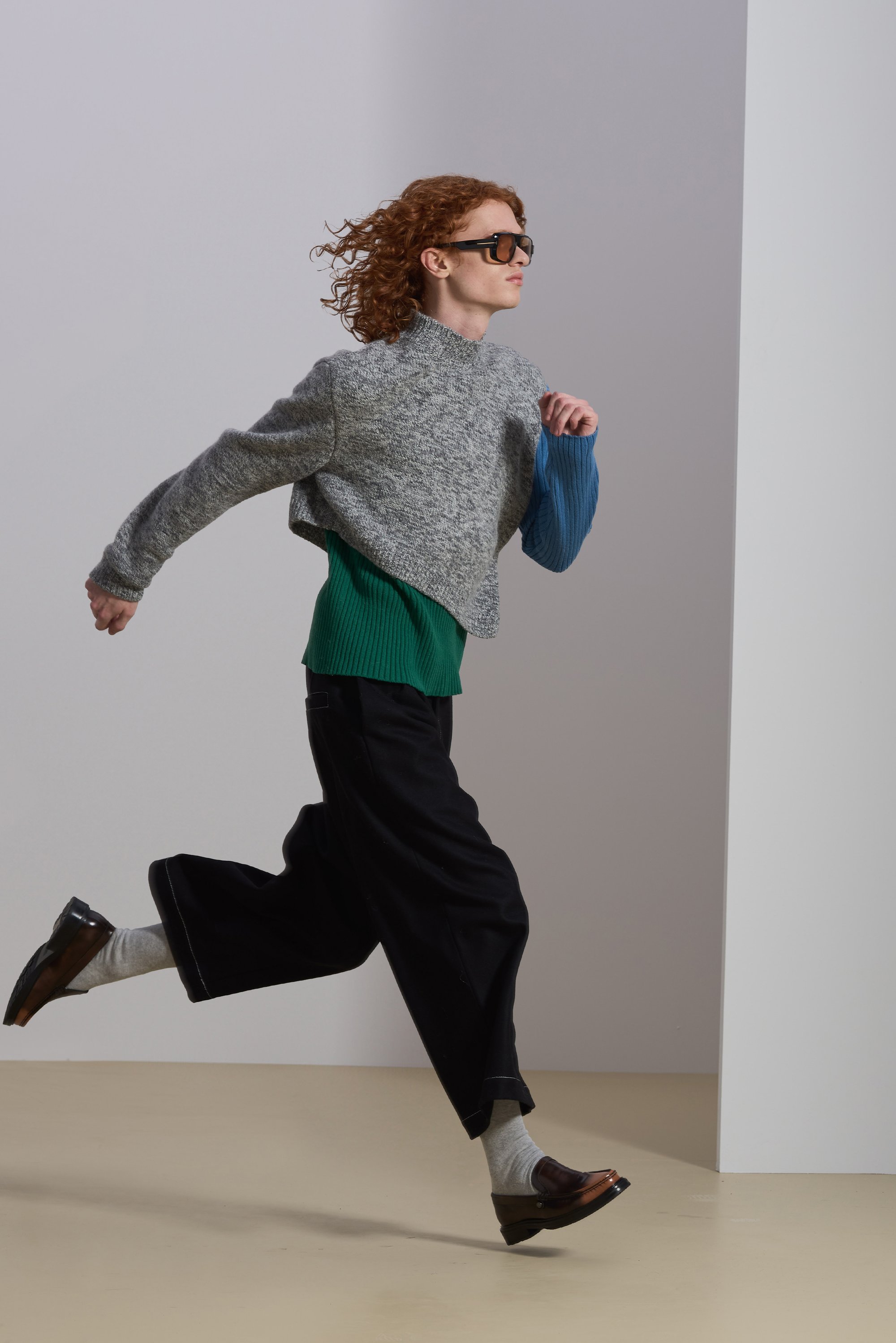 Light gray cropped sweater with a blue sleeve, paired with a green ribbed top and black wide-leg trousers, finished with brown shoes and gray socks.