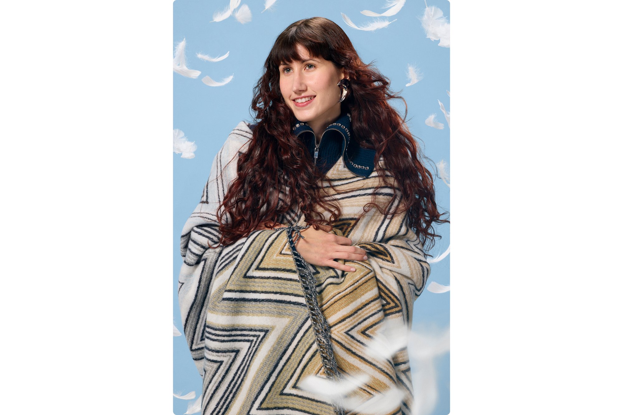 A woman with long, curly hair smiles while wrapped in a patterned poncho. White feathers float around her against a blue background.