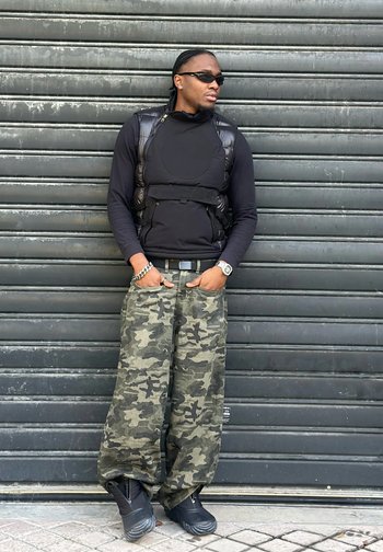 Hombre vestido con camiseta negra de manga larga, chaleco negro, pantalones anchos de camuflaje, gafas de sol y zapatos negros se apoya en una persiana metálica cerrada con las manos en los bolsillos.