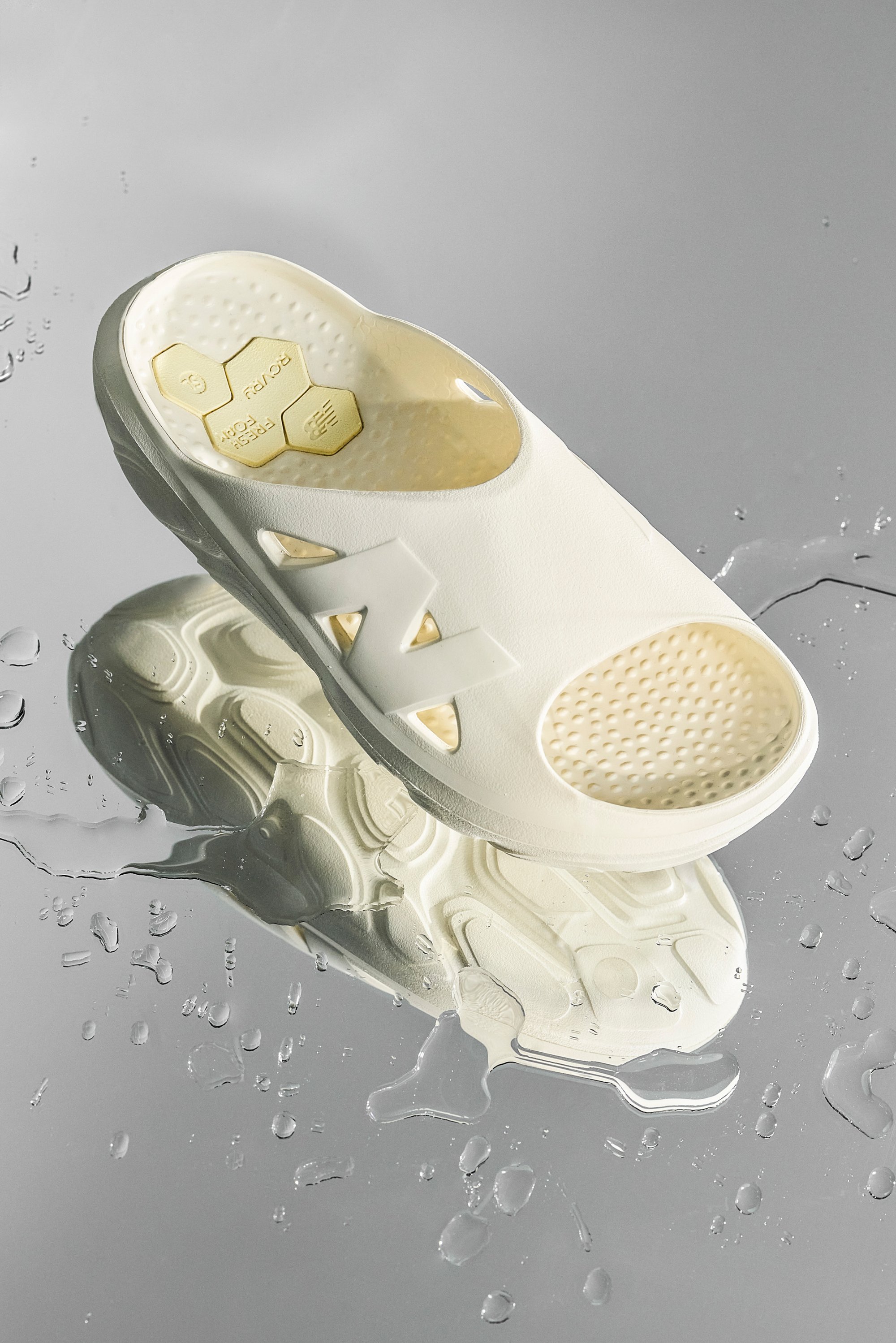 A white sandal rests on a reflective surface, surrounded by small water droplets, showcasing its textured sole and branding.