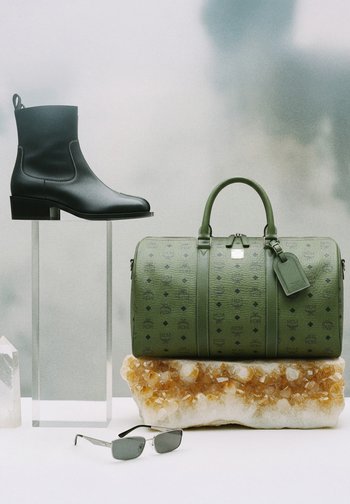 Mustast nahast pahkluu saabas läbipaistval alusel, rohelist mustriga reisikott merevaigu kristalli peal ning hõbedase raamiga päikeseprillid valgel pinnal.