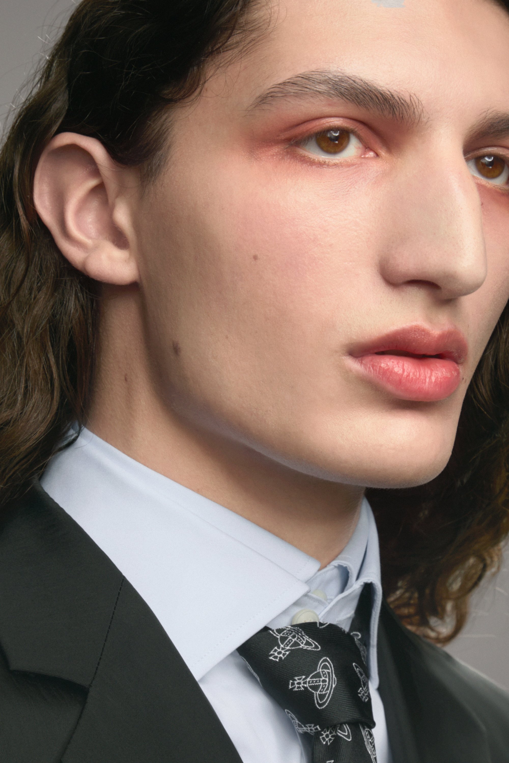 Una persona joven con cabello largo y oscuro viste un traje a medida y una corbata de patrón. Su maquillaje resalta sus ojos y labios, creando una apariencia impactante.