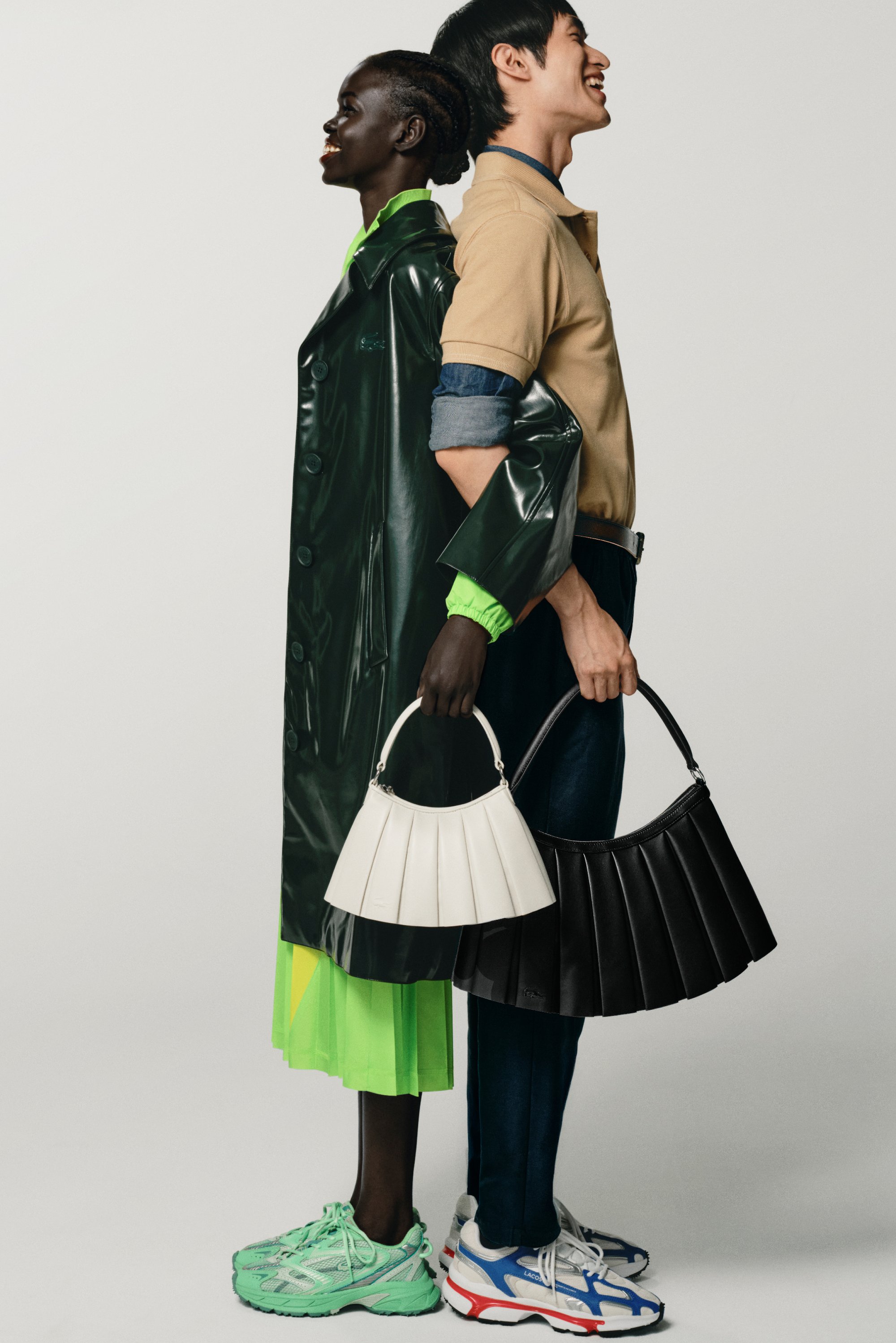 Cappotto lungo verde lucido, gonna verde neon, borse plissettate bianche e nere, scarpe sportive verde menta e blu con accenti rossi.