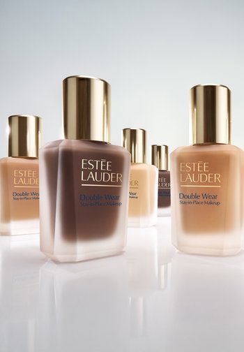 Cinq flacons de fond de teint Estée Lauder Double Wear Stay-in-Place Makeup dans différentes teintes, avec des bouchons dorés, sur une surface réfléchissante.