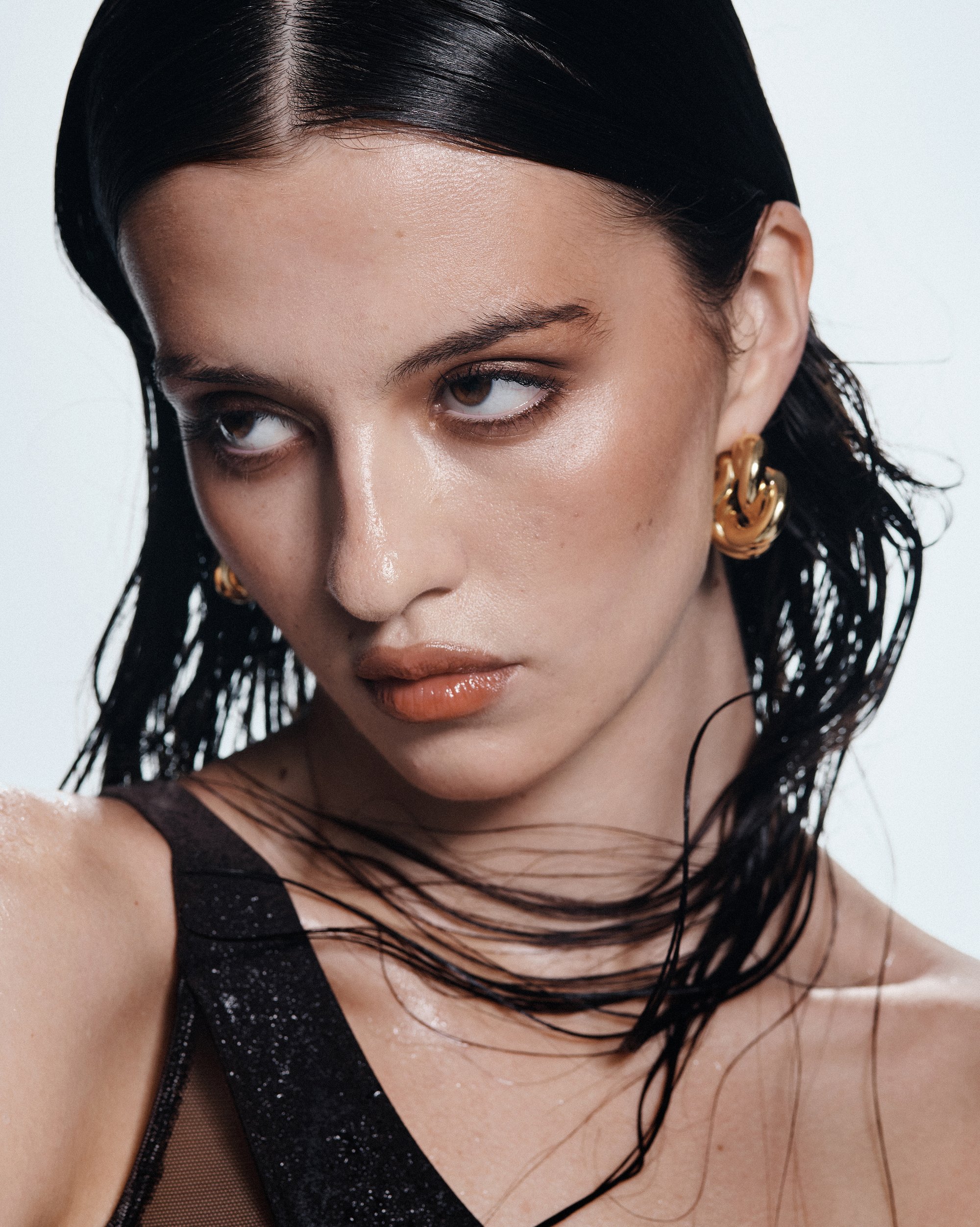 Een model met nat, donker haar toont een serieuze uitdrukking, draagt grote gouden oorbellen en een mouwloos, glinsterend topje tegen een lichte achtergrond.