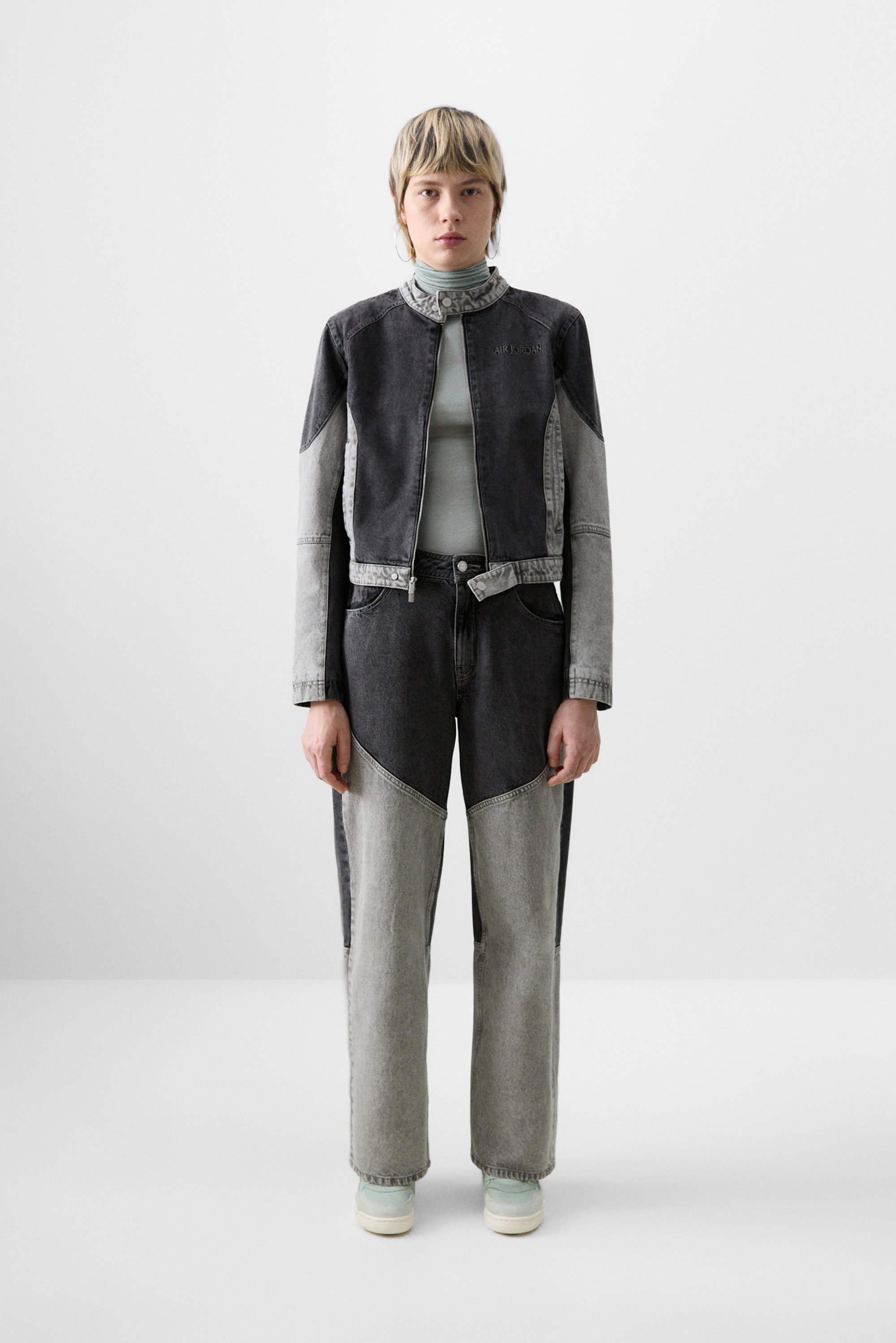 Una persona con capelli corti e chiari indossa una giacca di jeans cropped bicolore e pantaloni abbinati, in piedi contro uno sfondo bianco uniforme.