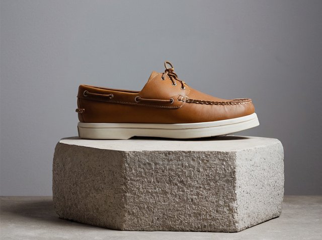 Braune Lederbootsschuhe mit weißer Gummisohle, ausgestattet mit dekorativen Nähten, Schnürsenkeln und metallischen Ösen, präsentiert auf einem strukturierten Betonpodest.