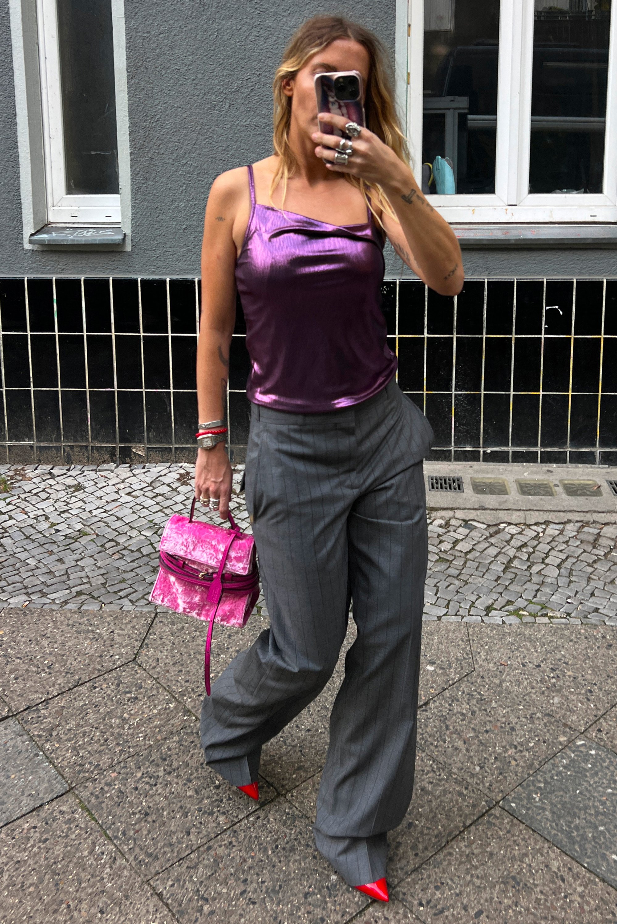 Camisola di raso viola, pantaloni a gamba larga grigio a righe, décolleté rossi brillanti e una borsa in velluto rosa con manico.
