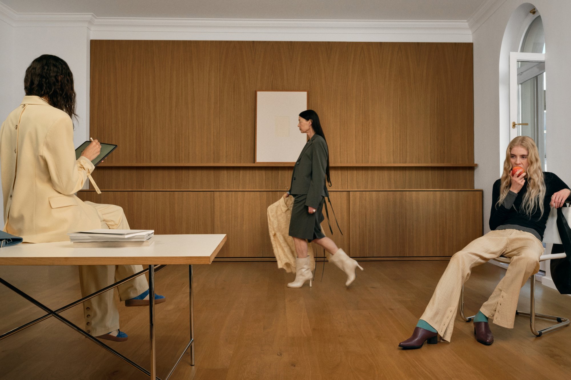 Tres mujeres en una habitación moderna y minimalista: una sentada escribiendo, otra caminando con una tela, y una comiendo una manzana, todas con atuendos elegantes.