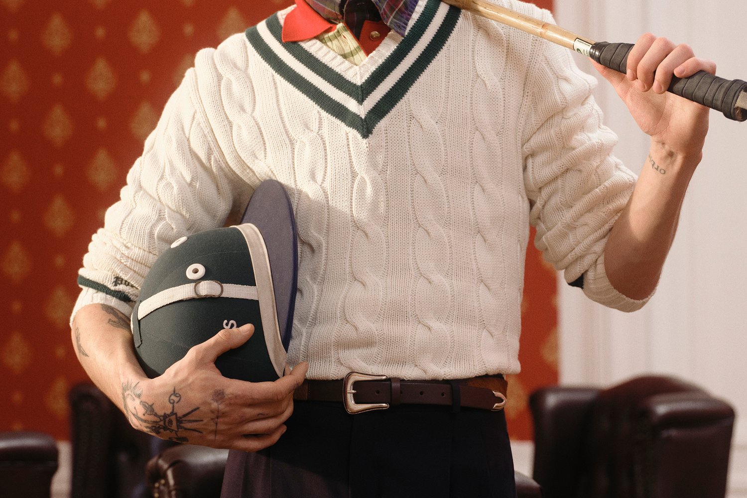 Persona con un suéter de punto trenzado sostiene un bate de cricket y un casco verde frente a una pared roja con patrones, mostrando un estilo deportivo clásico.