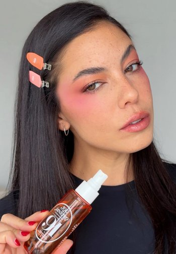 Frau mit pinkem Lidschatten und Haarklammern hält eine orange Sprühflasche der Sol de Janeiro 9-in-1 Haarbehandlung.