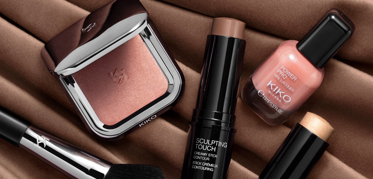 Compact blush, skulpturerende contour-stick, fersken neglelak, cremet highlighter og makeupbørste arrangeret på brun stof.