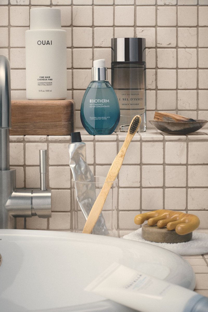 Bathroom sink area with a bamboo toothbrush, toothpaste tube, skincare bottles, soap, and a faucet against beige tiled wall.