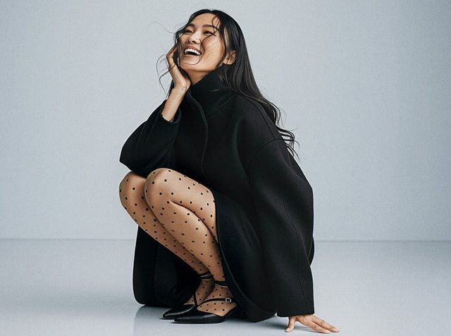 Oversized svart kappa, genomskinliga svarta strumpbyxor med prickar och spetsiga svarta klackar. Kappan har en hög krage och vida ärmar.