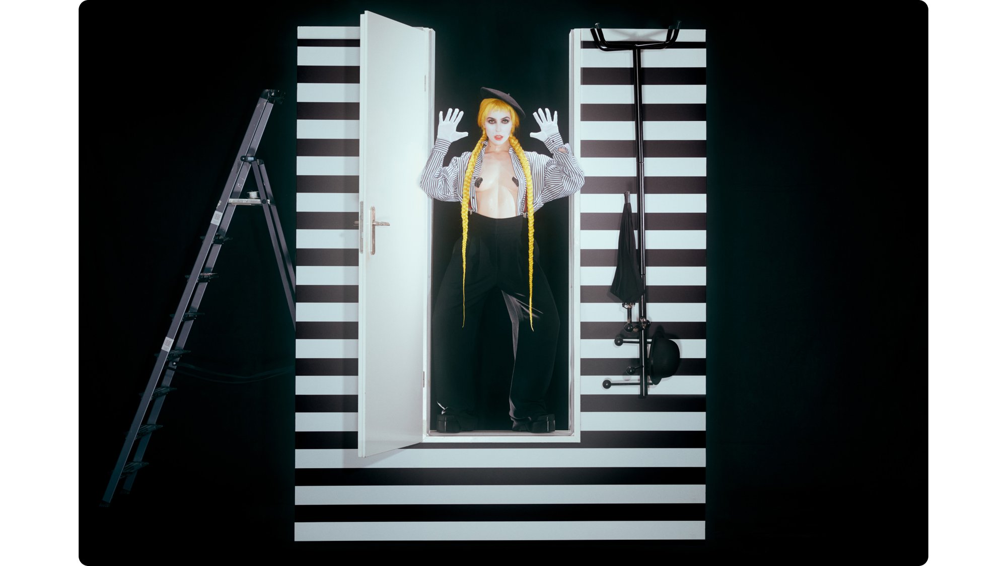 A person with bright yellow braids and striking makeup stands in an open doorway, hands raised, within a striped black and white background.
